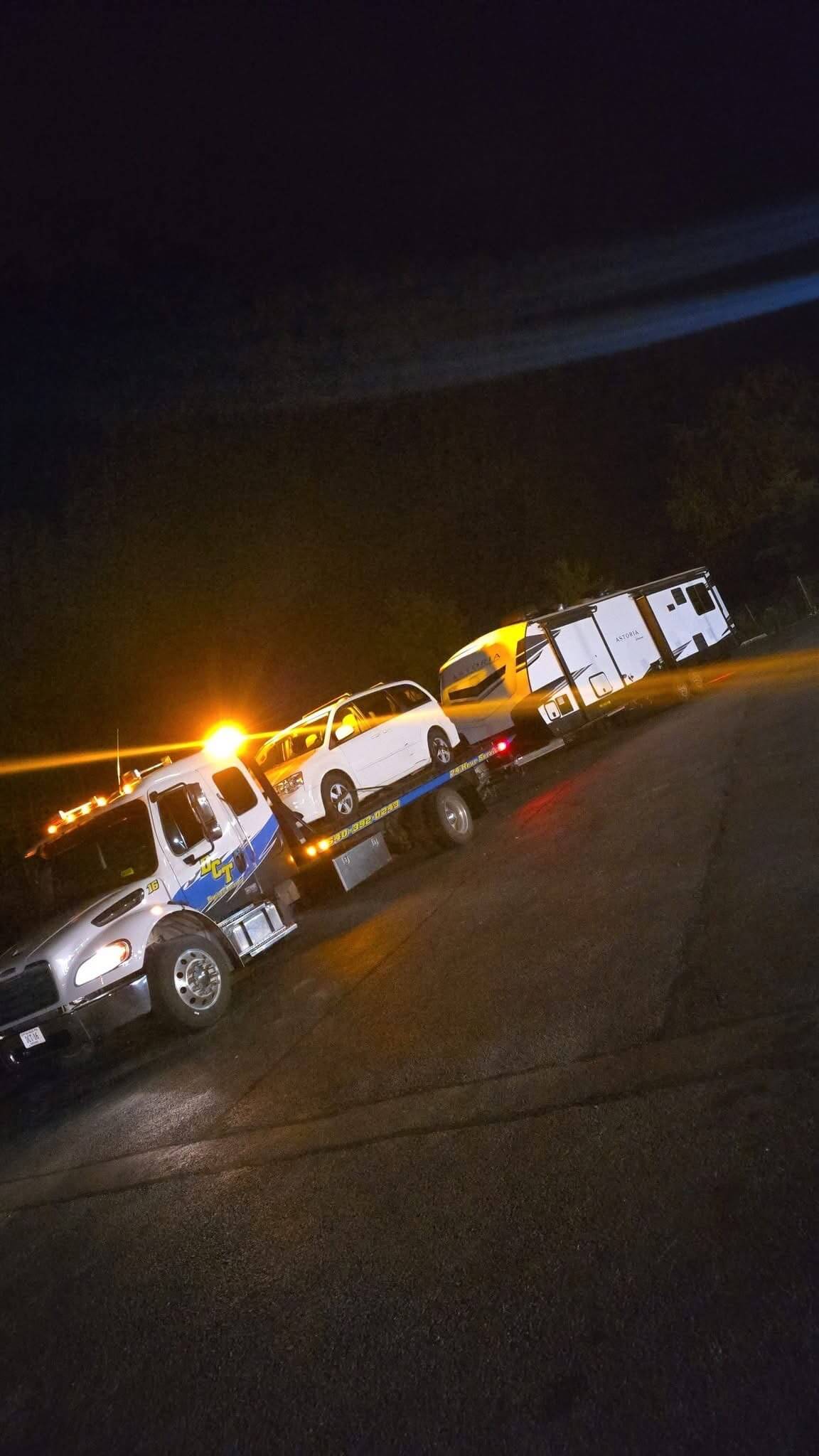 A DCT flatbed carrying a minivan while towing a travel trailer behind it at night.
