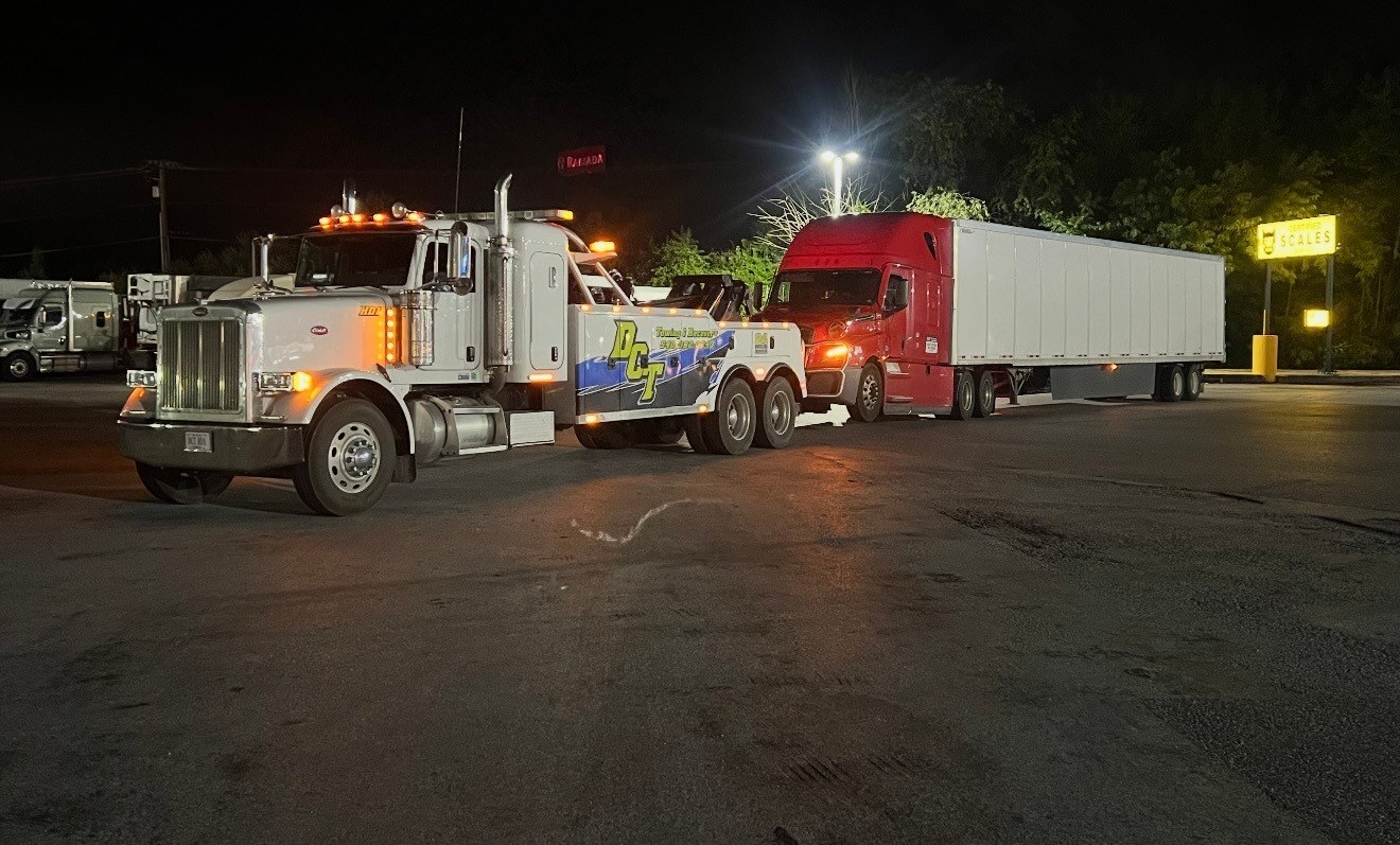 A DCT heavy wrecker hooked to a red tractor trailer at a truck stop at night.