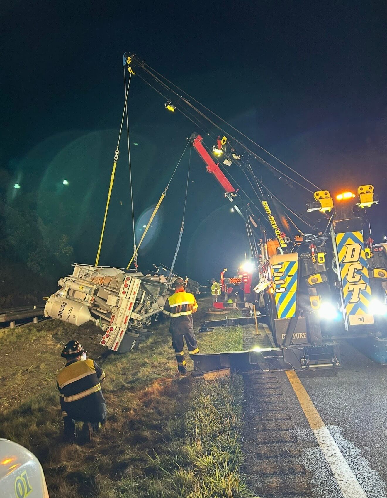 DCT crews in high visibility gear uprighting an overturned tanker trailer beside the interstate at night.