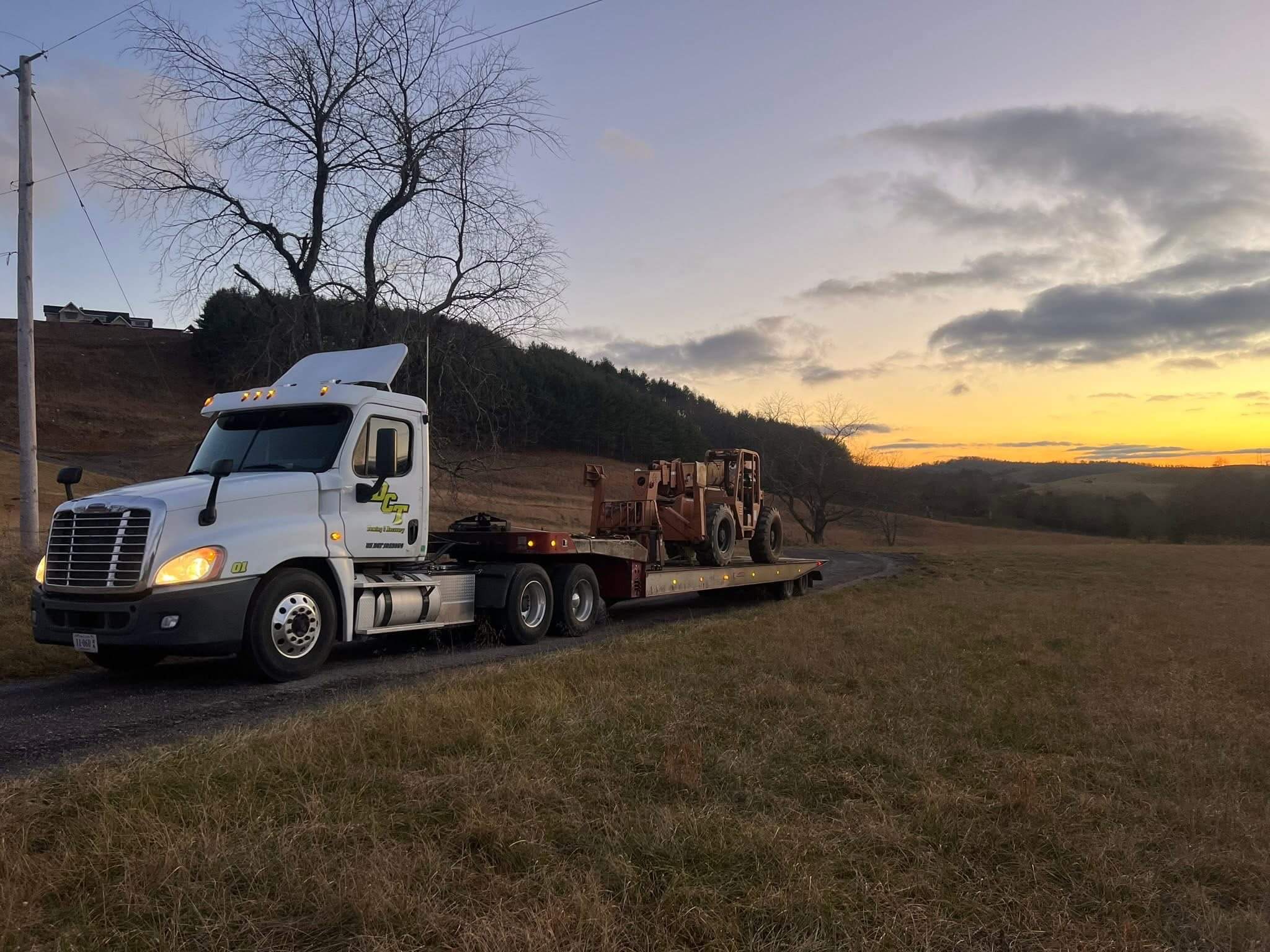 A DCT tractor and lowboy trailer hauling a rough terrain forklift down a country road at sunset.