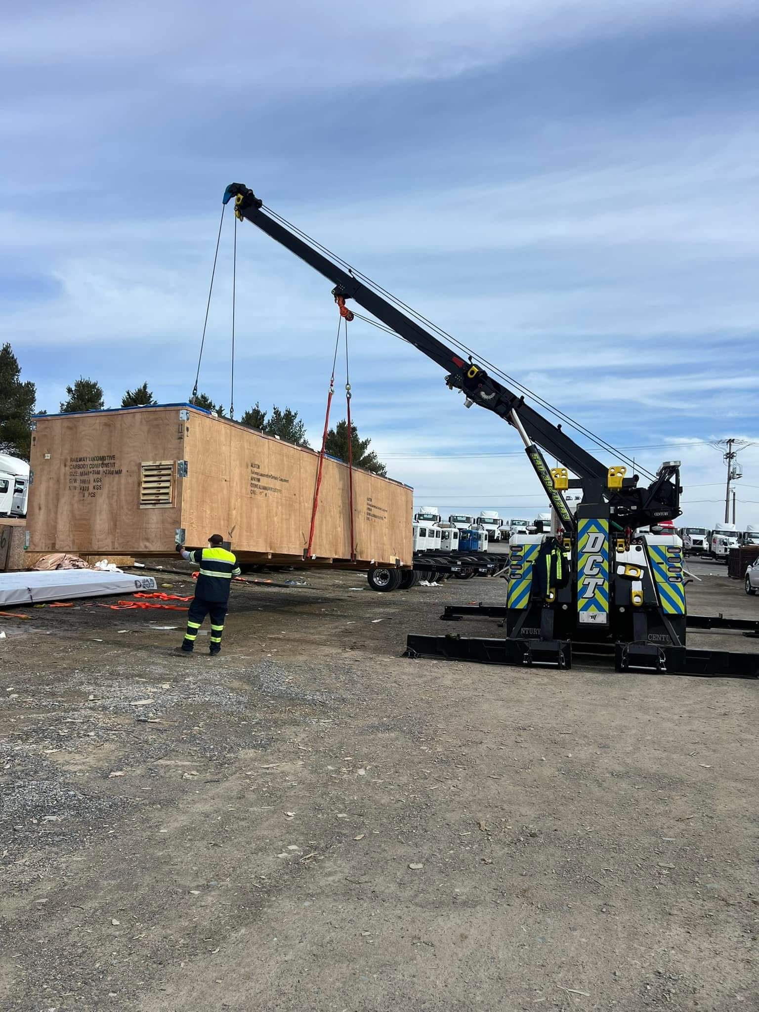 A DCT rotator wrecker lifting a large crated railway locomotive component at a freight yard.